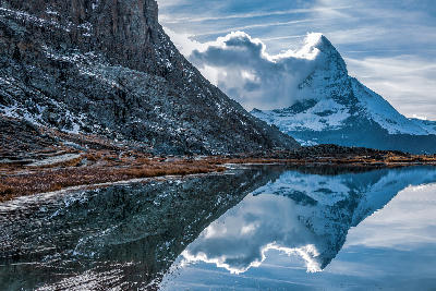 Matterhorn partially covered by clouds reflected in Riffelsee with alpine shoreline and rocky terrain, Zermatt, Switzerland.