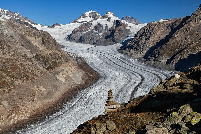 Aletsch Glacier flowing through a high alpine valley with rocky peaks and a stone cairn in the foreground, Switzerland.