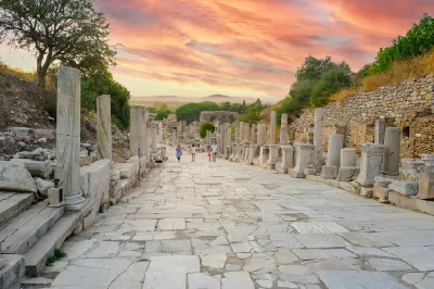 Marble-paved Curetes Street in Ephesus descending between rows of ancient columns and ruins beneath a colorful sunset sky.