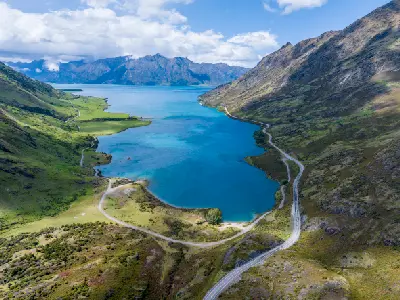 Aerial view of Lake Hāwea with winding lakeshore road and surrounding Southern Alps mountains, South Island, New Zealand.