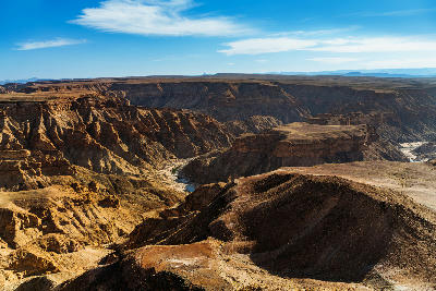 Wide elevated view across Fish River Canyon in Namibia showing layered canyon walls, plateau formations, and winding erosion channels beneath a blue desert sky.