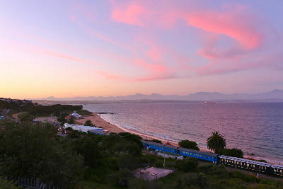 Sunset view of Santos Beach and the Santos Express Train Lodge in Mossel Bay with calm bay waters and mountain silhouettes in the distance.