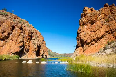 View looking through Glen Helen Gorge with rugged red-orange rock walls, a wide waterhole, and green grasses beneath a clear blue sky