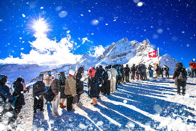 Tourists queuing at Jungfraujoch viewing area with Swiss flag and snow-covered peaks, Bernese Alps, Switzerland.