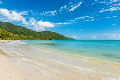Wide view from the sand looking across calm, clear turquoise ocean water towards forested tropical hills under a bright blue sky with wispy clouds.