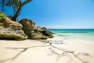 White sand, weathered coastal rocks and clear turquoise water framed by a tree at Murrays Beach in Booderee National Park.