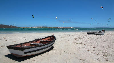 Fishing boats on the sandy shoreline of Langebaan Lagoon with kitesurfers on the turquoise waters near Langebaan, Western Cape, South Africa.