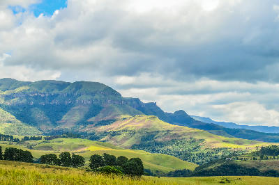 Green foothills and forested valleys beneath Champagne Castle, illuminated by patches of sunlight under dramatic storm clouds in the Central Drakensberg.