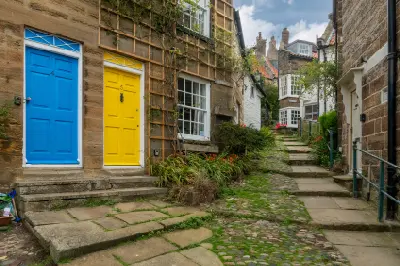 Bright blue and yellow cottage doors beside a cobbled passage lined with traditional stone houses and climbing plants in Robin Hood's Bay, North Yorkshire.