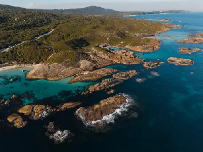 High-angle aerial view of vibrant turquoise ocean pools and sandy beaches framed by dramatic rocky reefs, rocky outcrops, and sweeping green coastal hills stretching out to the horizon.