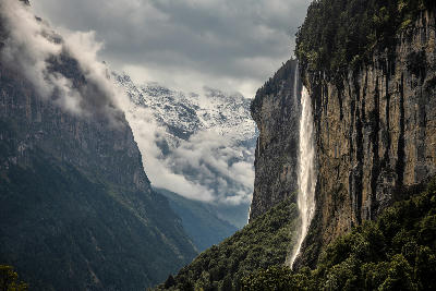 Staubbach Falls falling down a sheer rock face in Lauterbrunnen with forest below and snow-covered mountains partly hidden by clouds
