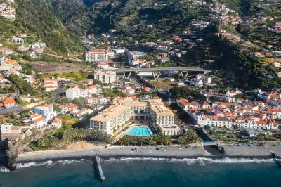 Aerial view of Santa Cruz in Madeira with beachfront hotel, palm trees, pebble beach, pier, and hillside houses overlooking the Atlantic Ocean.