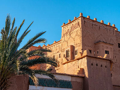 Detailed adobe architecture of Kasbah Taourirt in Ouarzazate with palm trees and geometric earthen wall patterns under a blue sky.