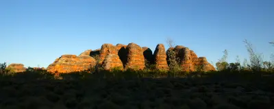 Warm evening light illuminates the distinctive striped sandstone domes of the Bungle Bungle Range above dark foreground bushland.