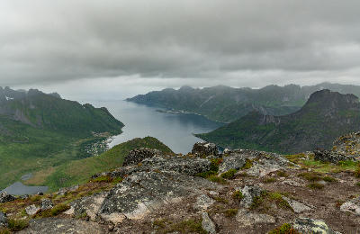 Elevated view from Barden showing rocky foreground, fjord basin, and Husøy village on a small island surrounded by steep mountains