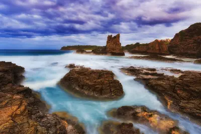 Cathedral Rocks rise from the turbulent ocean beneath dramatic storm clouds, with long-exposure waves flowing across the rocky shoreline.