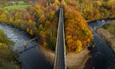 Top-down drone view along Lambley Viaduct, with the River South Tyne curving around golden autumn trees and a narrow footbridge visible below.