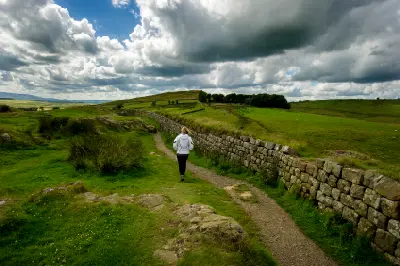 Walker on a footpath beside Hadrian's Wall at Steel Rigg, Northumberland, with green hills and large clouds overhead.