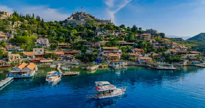 Boats cross the clear blue water in front of Kaleköy, where traditional red-roofed houses climb a green hillside toward the historic stone castle of Simena.