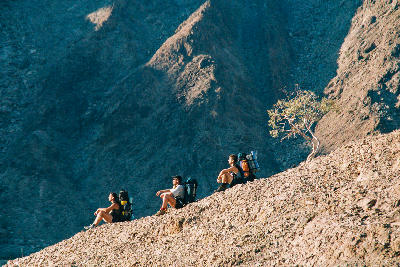Three backpackers resting on a rocky ridge inside Fish River Canyon in southern Namibia with steep canyon walls and arid desert terrain surrounding them.