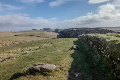 Hadrian’s Wall running beside a grassy walking path at Steel Rigg in Northumberland, England, with steep rocky cliffs extending into the distance beneath a partly cloudy sky.