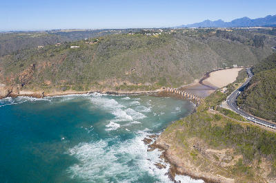 Aerial view of Kaaimans River Mouth near Wilderness, where a historic railway bridge crosses the estuary beneath steep coastal cliffs along South Africa's Garden Route.