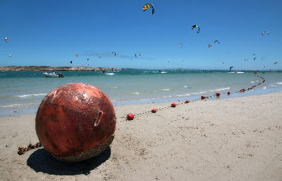Kitesurfers on Langebaan Lagoon with colourful kites above shallow turquoise water and a buoy line curving along a sandy beach on the West Coast of South Africa.