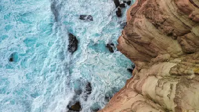 Turquoise waves break against layered sandstone cliffs around Natural Bridge.