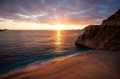 Golden sunset over Kaputaş Beach, with waves washing onto the sandy shore beneath a steep rocky headland and warm light reflecting across the Mediterranean Sea.