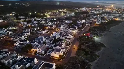 Aerial dusk view of Paternoster showing whitewashed houses, glowing streetlights and beachfront roads beside the Atlantic Ocean on South Africa’s West Coast.