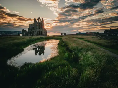 Gothic ruins of Whitby Abbey silhouetted against an orange sunset sky, with reflections in a foreground pool on the East Cliff in North Yorkshire, England.