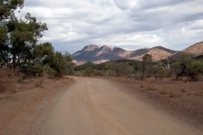 Unsealed Moralana Scenic Drive curving through dry eucalyptus bushland towards rugged mountain peaks beneath an overcast sky in South Australia's Flinders Ranges.
