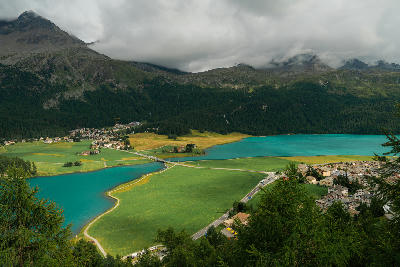 Elevated view of Lake Silvaplana with turquoise water, village, fields, and forested slopes beneath low clouds in the Engadin, Switzerland