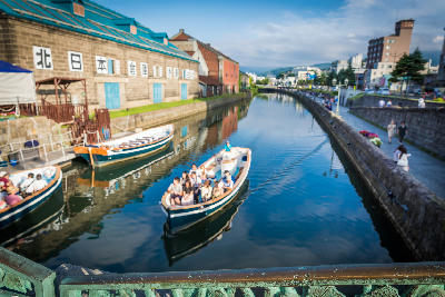 Boat carrying visitors along Otaru Canal with historic warehouse buildings and reflections in Hokkaido, Japan.