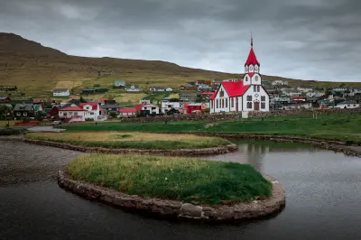 Sandavágur Church with red roof beside a small pond, with village houses and grassy hills behind under overcast skies on Vágar Island, Faroe Islands.