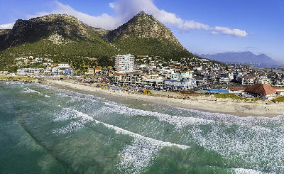 Aerial view of Muizenberg Beach with surfers, sandy shoreline, beachfront buildings, and mountains behind on the False Bay coast in Cape Town, South Africa.