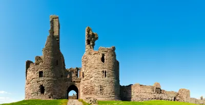 Close-up of Dunstanburgh Castle's ruined medieval gatehouse, showing two partially collapsed stone towers, an arched entrance and adjoining defensive walls beneath a clear blue sky.