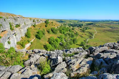 Limestone rocks in the foreground overlooking the cliffs of Malham Cove, with green fields, dry-stone walls and rolling hills in the Yorkshire Dales.