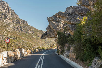 Mountain road passing beneath the rocky overhang of Dacres Pulpit in Bainskloof Pass, South Africa, surrounded by steep cliff faces and rugged mountain terrain.