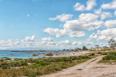 Dirt road leading to a jetty on the shoreline of St Helena Bay, with coastal vegetation, rocky outcrops, blue ocean and scattered clouds above South Africa’s West Coast.