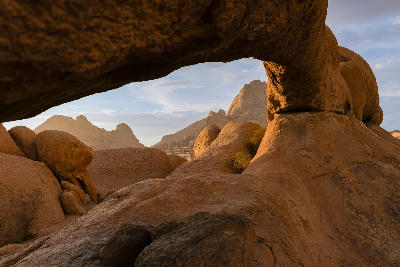 View through a granite rock arch at Spitzkoppe toward desert mountain formations in warm sunrise light.