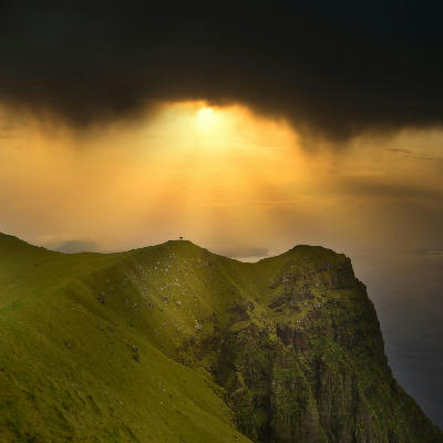 Kallur Lighthouse on a narrow grassy ridge at the edge of Kalsoy Island under dramatic storm clouds in the Northern Islands of the Faroe Islands.