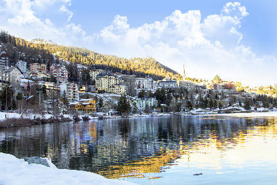 St. Moritz seen from Lake St. Moritz with winter light reflecting on the water, showing lakeside buildings, snow patches, and forested slopes in the Upper Engadin, Switzerland.