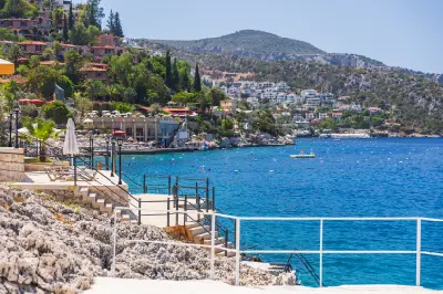 Rocky waterfront terraces and sunbathing areas line the vivid blue bay of Kalkan, with lush hillside villas rising toward rugged Mediterranean mountains.