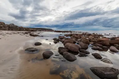 Rounded rocks and reflective tidal pools along Stokes Bay Beach on Kangaroo Island, beneath dramatic grey clouds.