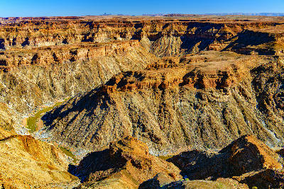 High elevated view across Fish River Canyon in Namibia showing steep layered canyon walls, rugged desert formations, and the winding river below beneath a clear blue sky.