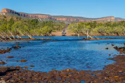 Vehicles approach the shallow Pentecost River crossing with rocky water in the foreground and the red escarpments of the Cockburn Range beyond.
