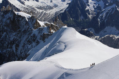 Climbers traversing a narrow snow ridge with steep drops in the Mont Blanc Massif, surrounded by high alpine peaks and glaciers.