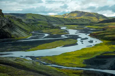 Meandering river flowing through the Icelandic Highlands, cutting through black volcanic sand and moss-covered hills under diffused light.