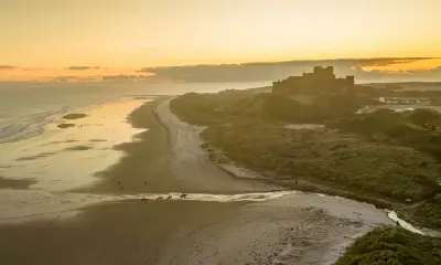 Aerial view of four racehorses crossing Bamburgh Beach at dawn, with Bamburgh Castle silhouetted above the sandy coastline beneath a golden sky.